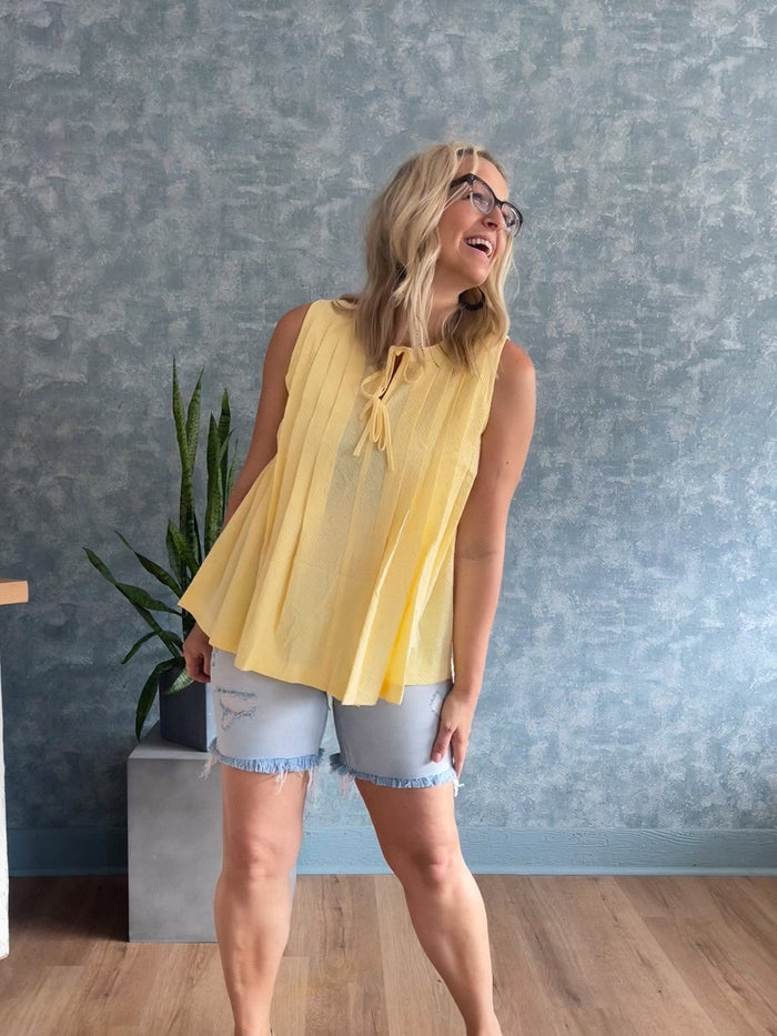 Woman wearing a yellow sleeveless top and denim shorts standing in a room with a gray wall and wooden floor.
