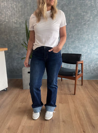 Person wearing a white shirt and blue jeans standing in a room with a chair and plant.