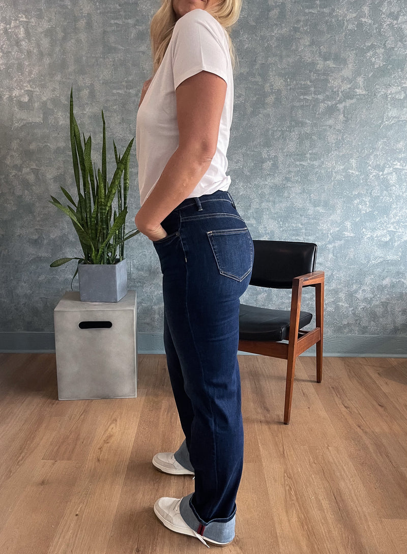 Person wearing blue jeans and a white shirt standing in a room with a plant and chair.