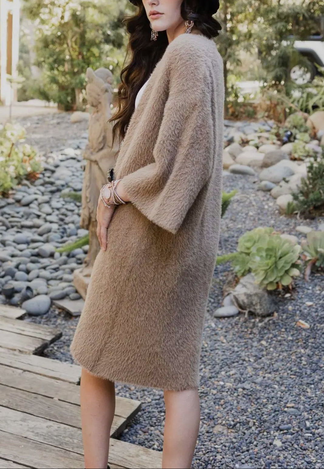 Woman wearing a long beige cardigan and black hat in a garden setting